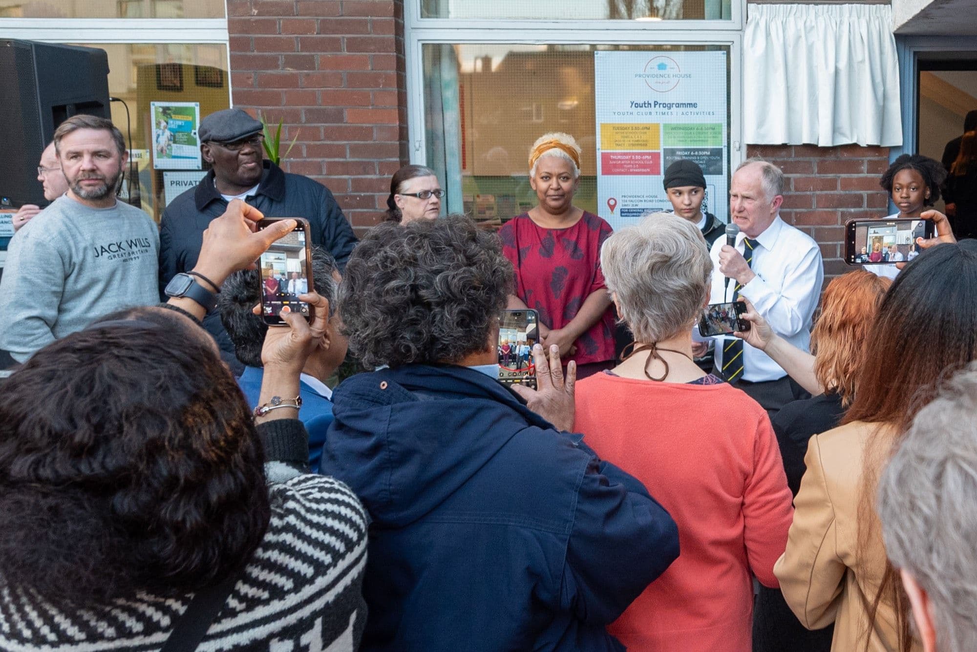 Neighbours, young people and supporters gather outside the Providence House entrance on Falcon Road as a speaker addresses them, many filming on their phones